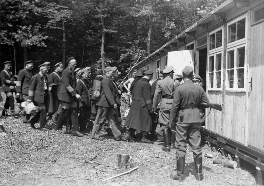 Unter Bewachung von SS-Angehörigen werden die ersten Schutzhäftlinge in eine der fertiggestellten Holzbaracken des Lagers gebracht, 15. Juli 1937
