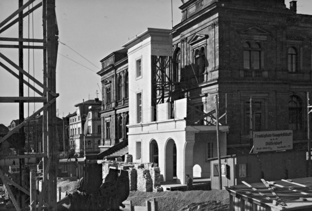 Fassadenmodell für das »Haus der Gliederungen der NSDAP« vor dem Landesmuseum, errichtet im September 1937, im Vordergrund werden durch eine Spezialfirma aufwendige Pfahlgründungen ausgeführt, bevor bis Ende Dezember die Maurerarbeiten bis zur Höhe der Kellerdecke beendet werden konnten